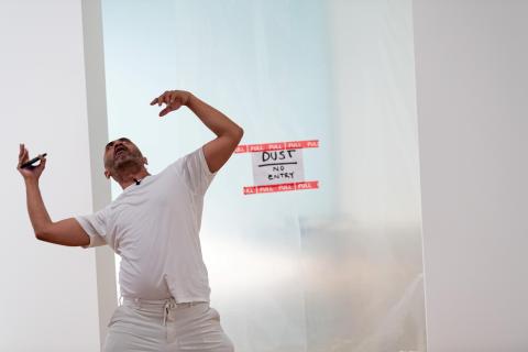 A man dressed in white dances with his hands in the air in front of a sign that warns 'no entry' due to dust.
