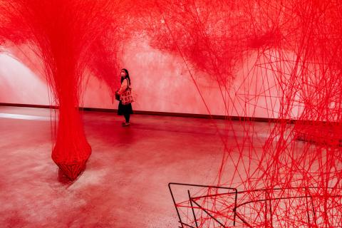 An installation view of a room completely webbed in bright red thread, some of which is tied down to metal boat structures on the gallery floor. A visitor walks through this red space.