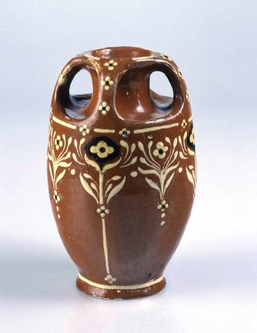 Artwork Vase this artwork made of Earthenware, hand-built white clay with four handles extending from lip to shoulder.  Dipped Redcliffe clay slip with formalised flower pattern in cream (Stradbroke Island) clay and black stained clay slip.  Clear glaze, created in 1931-01-01