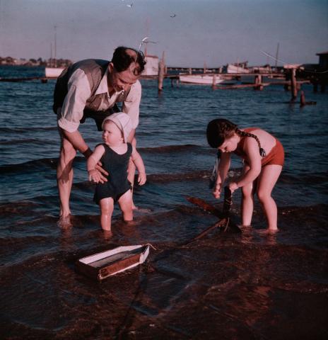 Artwork (J.H. Simmonds, Margaret and Rosemary standing in shallow water with toy boat) this artwork made of Cellulose acetate Dufay colour transparency (originally in a glass mount), created in 1935-01-01