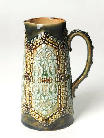 Artwork Jug this artwork made of Stoneware tapering cylindrical jug with elongated sprigged hexagonal design with gillouche border and daisy bosses enclosing foliate motifs.  Daisy bosses on handle.  Salt glazed green, light green and brown, created in 1884-01-01