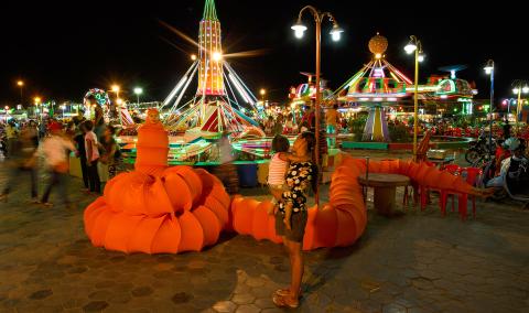 A still photograph taken at night in a fun fair; the artist is costumed as a red caterpillar-like bug with a very long body and tail.