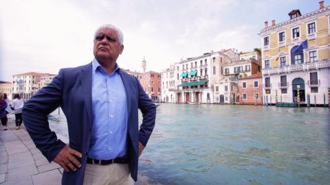 A still photograph of a man in a blue suit jacket standing in front of a canal in Venice.