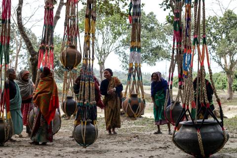 Women in brightly coloured clothes gather around large pots they have made, suspended from brightly coloured woven ropes.