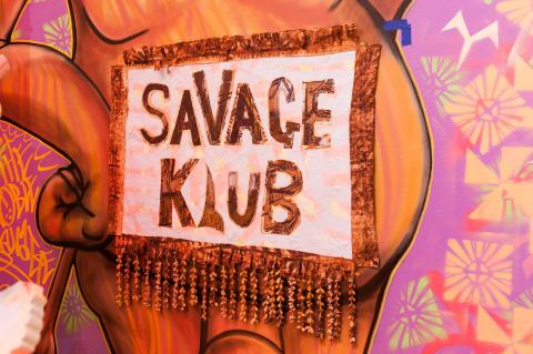 An installation view of a sign that reads 'SAVAGE KLUB' in chunky letters; its background is a brown and pink mural.