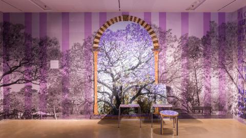 An installation view of a room with purple striped wallpaper and jacaranda tree accents.