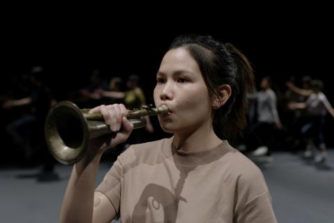 A still photograph of a young woman in a t-shirt playing a bugel.