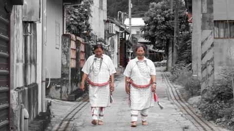 A still photograph of two Taiwanese women wearing traditional garments, walking down a road. The background is in black-and-white while the women are in colour.