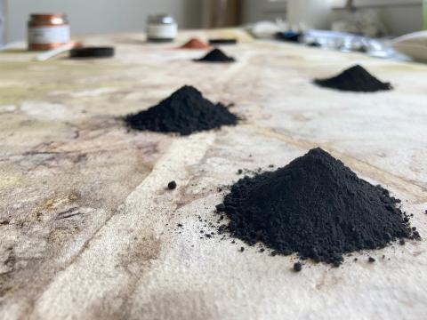 Small piles of black pigment on a fabric surface, with small pots in the background.