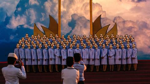 A digital photograph depicting a group of women in purple skirt-suits arranged for a photograph; they each have identical gold mask-like faces. A cloudy blue sky is behind them. Three people stand in the foreground taking photos.