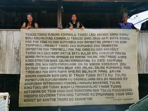 Three women on a balcony hold a mat over the side; the mat is woven with text in block letters.