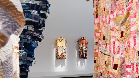 An installation view of works made of bright fabric in the foreground, with two garments hung against a white wall in the background; these are dresses with scenes embroidered on them.
