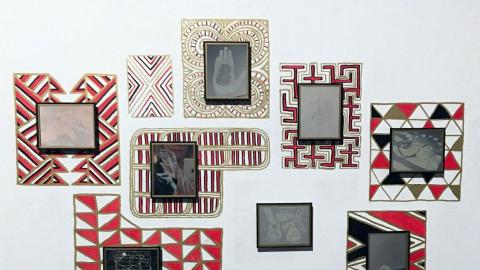 An installation view of small works hung salon-style on a gallery wall, with borders of bold black and red geometric patterns around each frame.