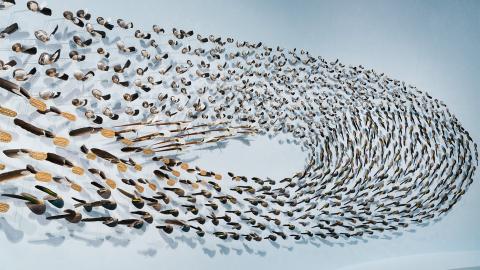 A detail view of an installation made of feathers and tools swirling in a wave-like formation against a light blue wall.