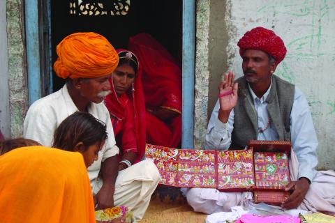 A storyteller (at right) holds a painted Kaavad, which he uses as a prompt; he performs for a group of people, of varying ages, who sit at left.