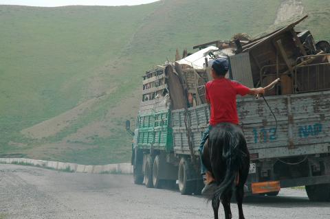 A still photograph o a track travelling through a mountainous landscape; behind the truck, a person dressed in red rides a horse.