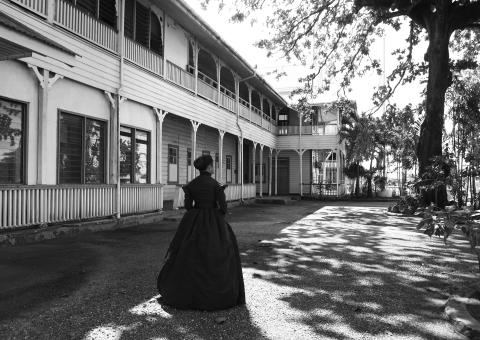 A black-and-white photograph of a large wooden colonial building; a Samoan woman stands out front, facing away from the camera, wearing 19th-century garb.