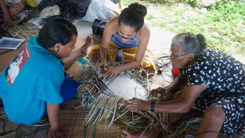 A group of women sitting together on the ground, weaving.