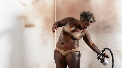 A photograph of a performance artist dressed in underwear, using a spray-gun to apply artificial colouring solution to her skin (much like self-tanning).
