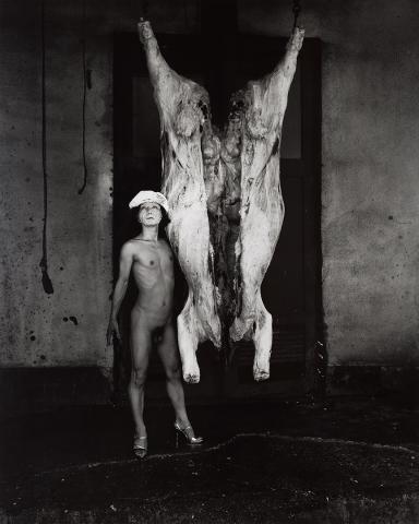 A black-and-white photograph of a naked man wearing a beret and high heels standing next to a flayed bovine carcass.