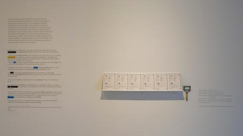 An installation view of a work featuring text on a white wall and a shelf with documents and a magnifying glass.