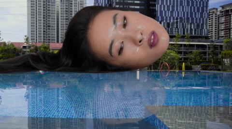 An enormous woman's head floats over a swimming pool. She has glossy, wet skin and long hair and looks towards the pool.