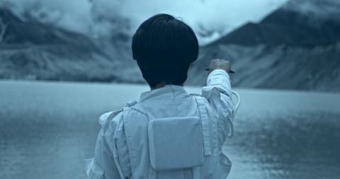 A person with short hair and wearing futuristic clothes faces away from the camera and points towards a lake.