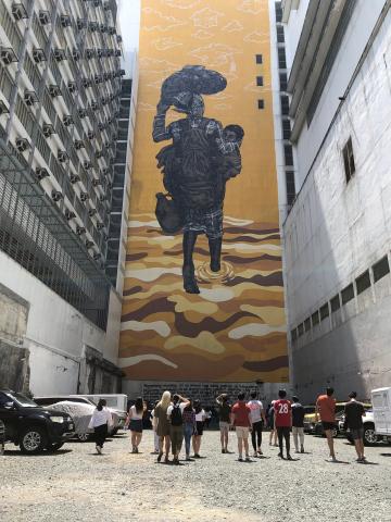 Tall mural of a woman walking through water while holding a child, painted on the side of a building. A crowd of people below looks up.