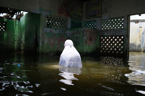 A photo of a person dressed in a white robe and head covering standing elbows deep in water inside a flooded building. They face away from the camera.