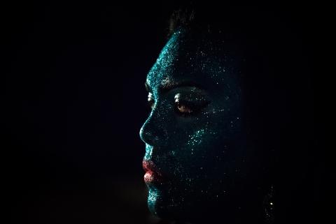 A still photo with a jet-black background; the face of a person with teal glitter covering their entire face, except red lips, emerges from this darkness.