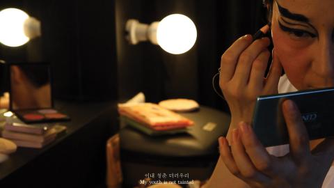 A still photograph of a person putting on make-up; they sit at a mirror with lightbulbs around it.