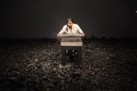 A photograph of a woman standing in the middle of an art installation, wherein she crushes and grinds hundreds of kilograms of charcoal briquettes into powder and dust.