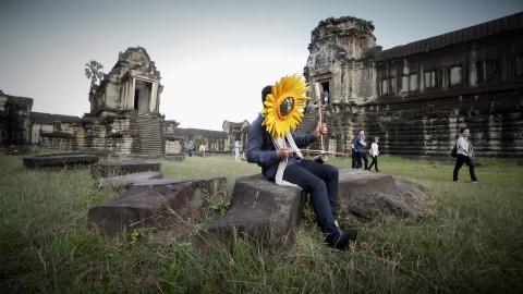 A man wearing a sunflower masks plays music amongst ruins.