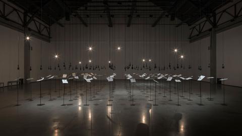Installation view of many pieces of paper, each speared on a sharp point, surrounded by light bulbs and speakers in a dark room.