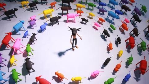 A still from a stop-motion video, in which a minotaur is surrounded by concentric circles of brightly coloured cows and calves.