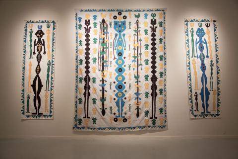 An installation view of three works on cotton; the middle of the three is the largest, and each has a stylistic human figure in the centre, surrounded by bright geometric and animal shapes