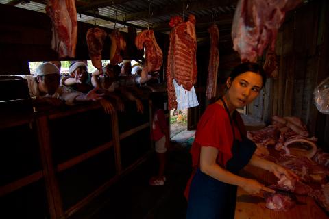A photograph of a woman working in a butcher shop surrounded by carcasses and pieces of meat; behind her, men reach in through a narrow window.