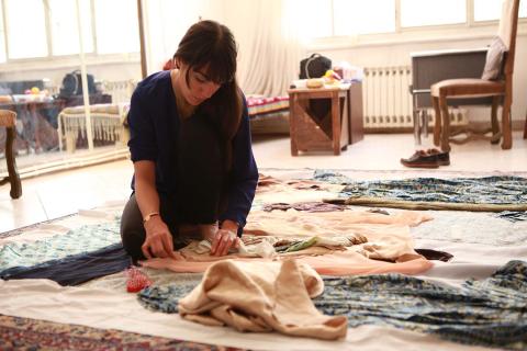 A woman in a well-lit room sits on the floor working on a textile-based artwork.