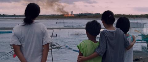 A photograph of four children, their backs to the camera, watching a boat in the far distance, on fire in the water.