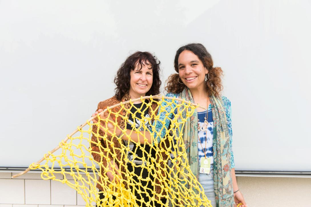 Two women stand in front of GOMA's external wall holding a yellow fishing net and smiling.