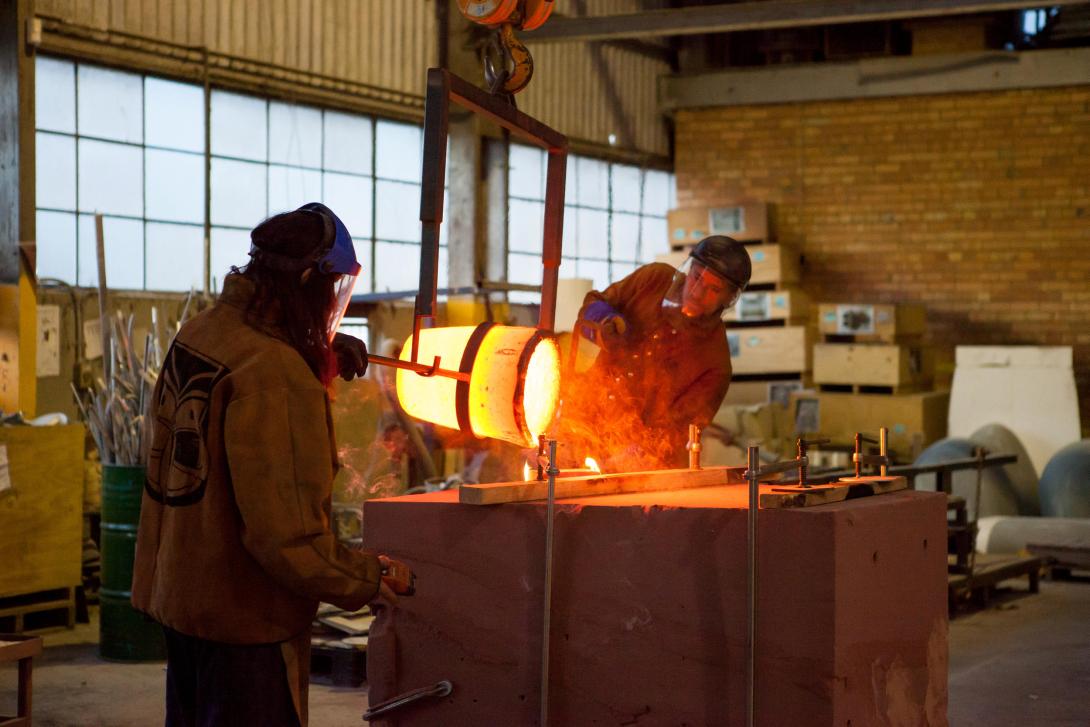 Two workers pour moulten bronze at a foundry.