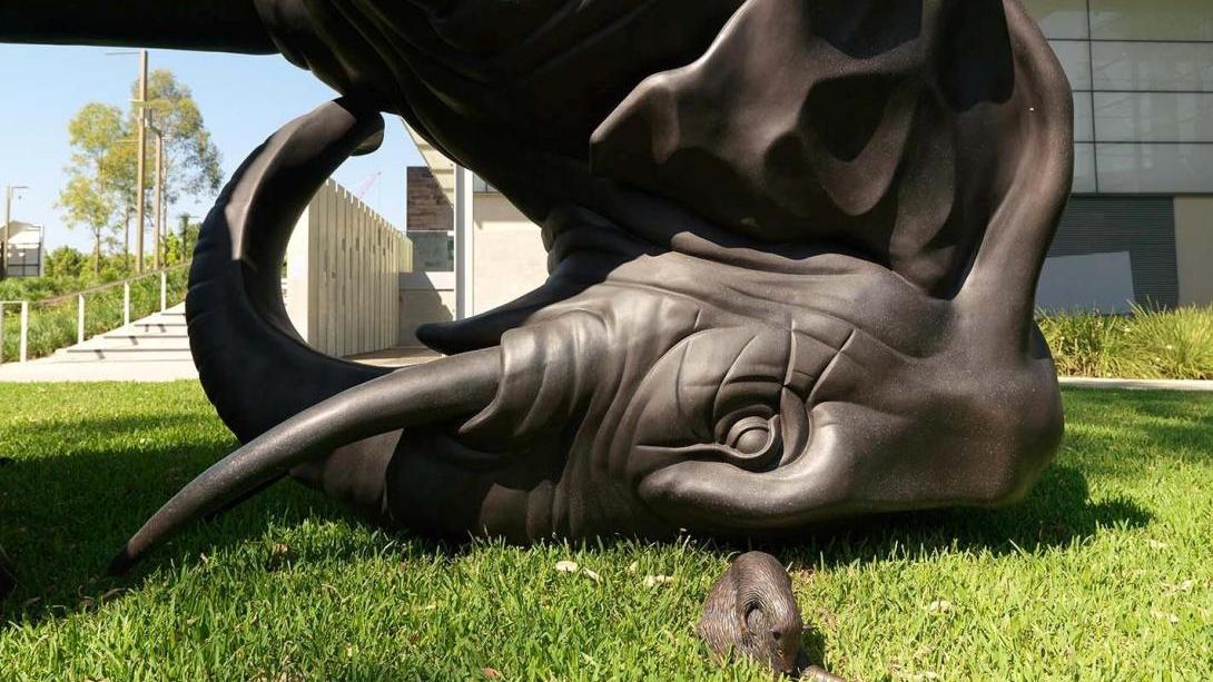 An installation view of a bronze sculpture set on grass outside GOMA, depicting an upside-down elephant eye-to-eye with a water rat.