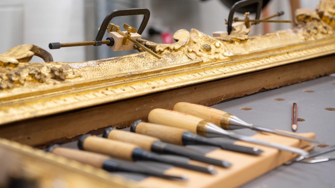 A selection of carving and woodworking tools on a table, set beside a gold frame in the process of being restored.