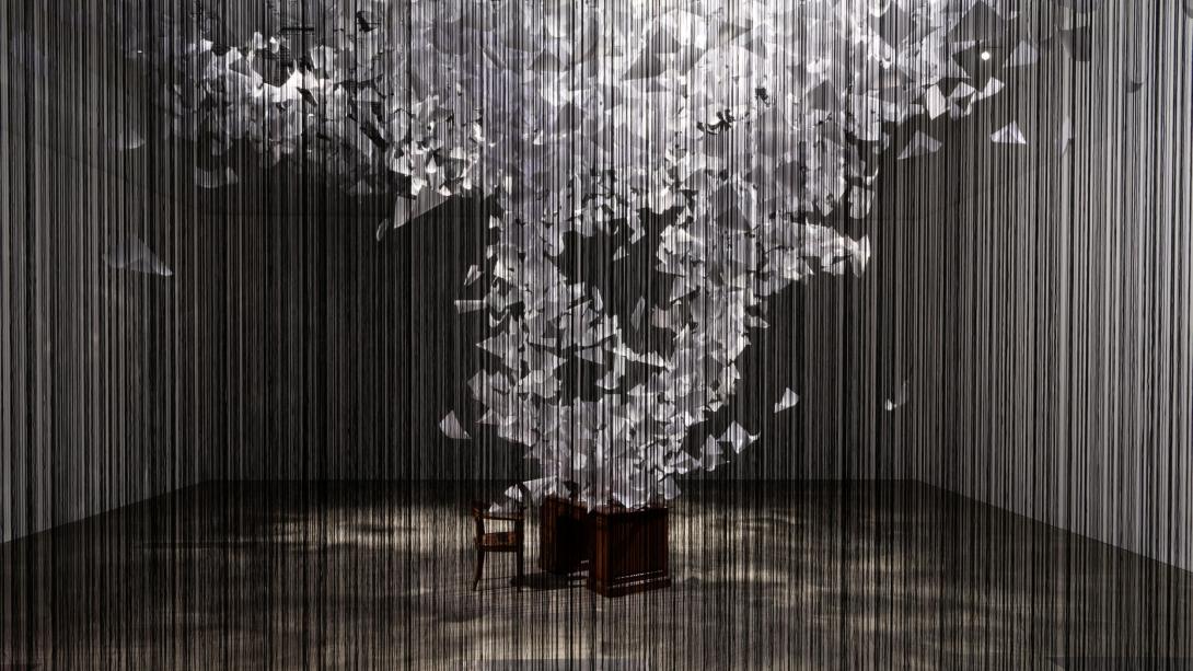 An installation view of a work that fills a whole room; at the centre, a desk and empty chair have thousands of papers appearing to fly off of them in a spiral. Suspended around this scene is a web of black threads.