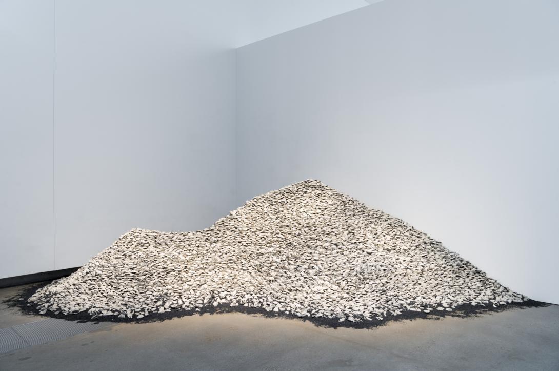 An installation view that appears as a large midden of white shells piled in front of a white gallery wall, in a corner.