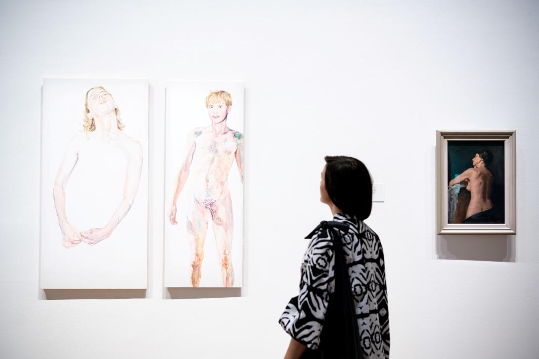 An installation view of a gallery visitor looking at two self-portraits by Tyza Hart, with another smaller self-portrait by Gwendoline Grant at right, installed on a white gallery wall.