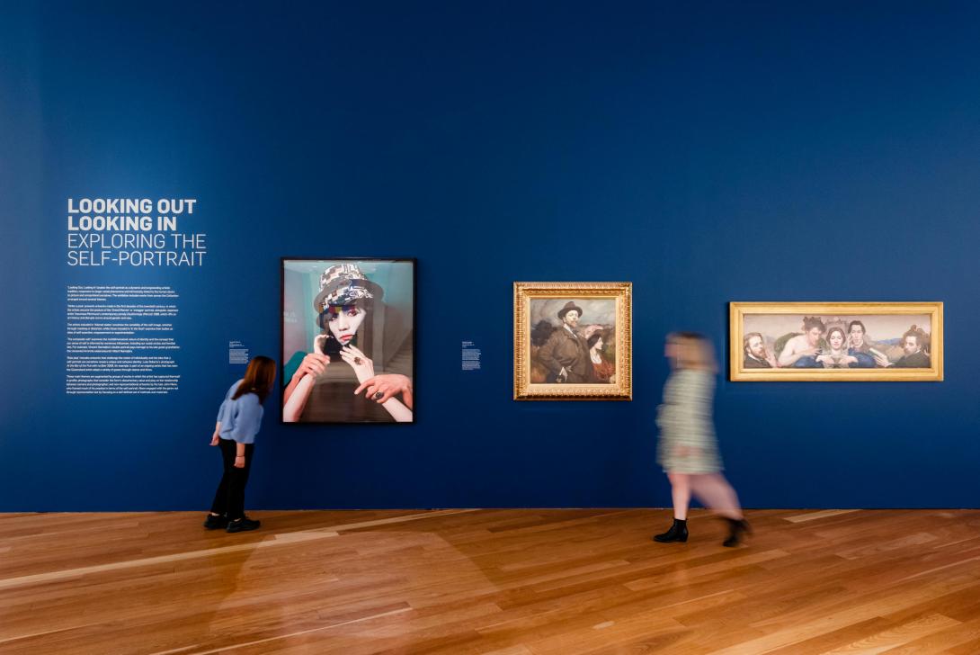 An installation view of three bold swagger portraits hung on a deep blue wall, with visitors walking by on the polished wood floor.