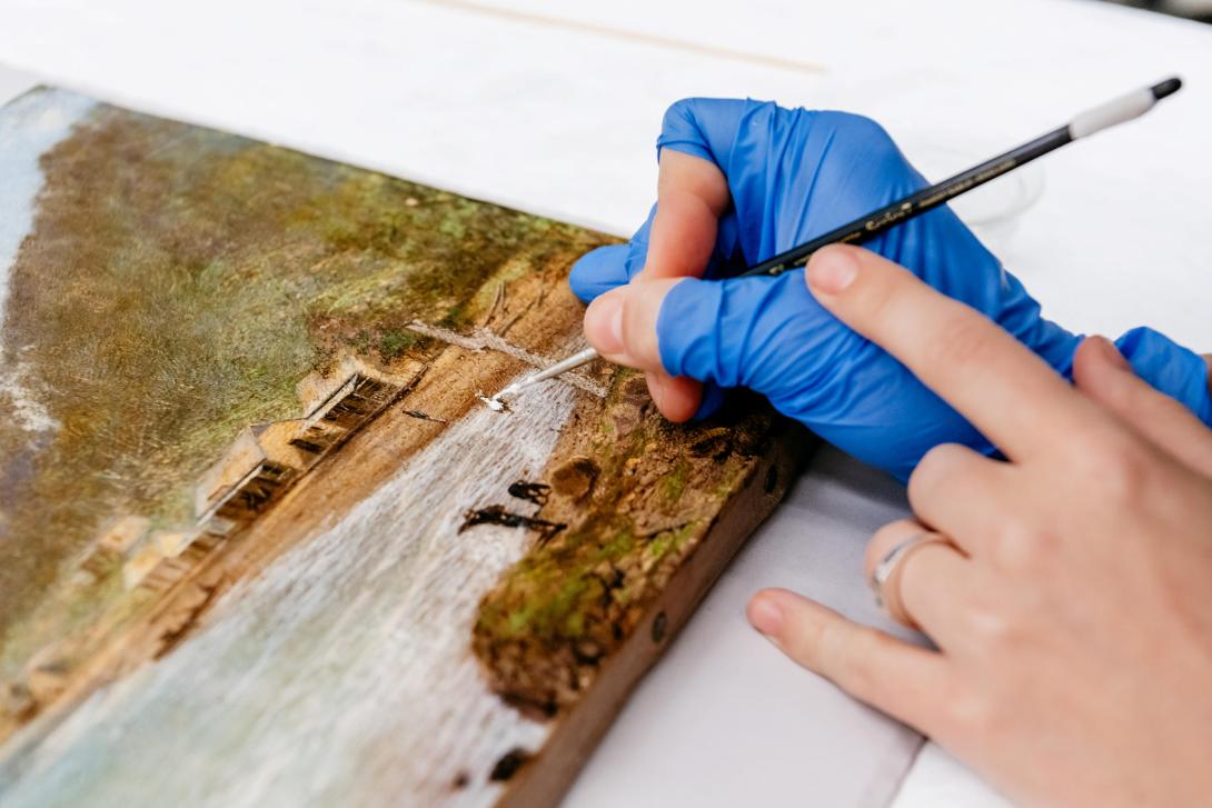 In a close-up photograph, the hand of a conservator performs restoration work with a small paintbrush; the conservator wears fingerless blue latex gloves.