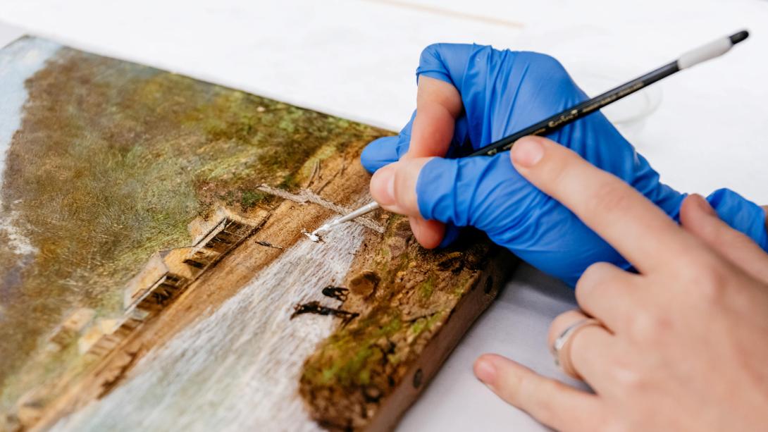 In a close-up photograph, the hand of a conservator performs restoration work with a small paintbrush; the conservator wears fingerless blue latex gloves.