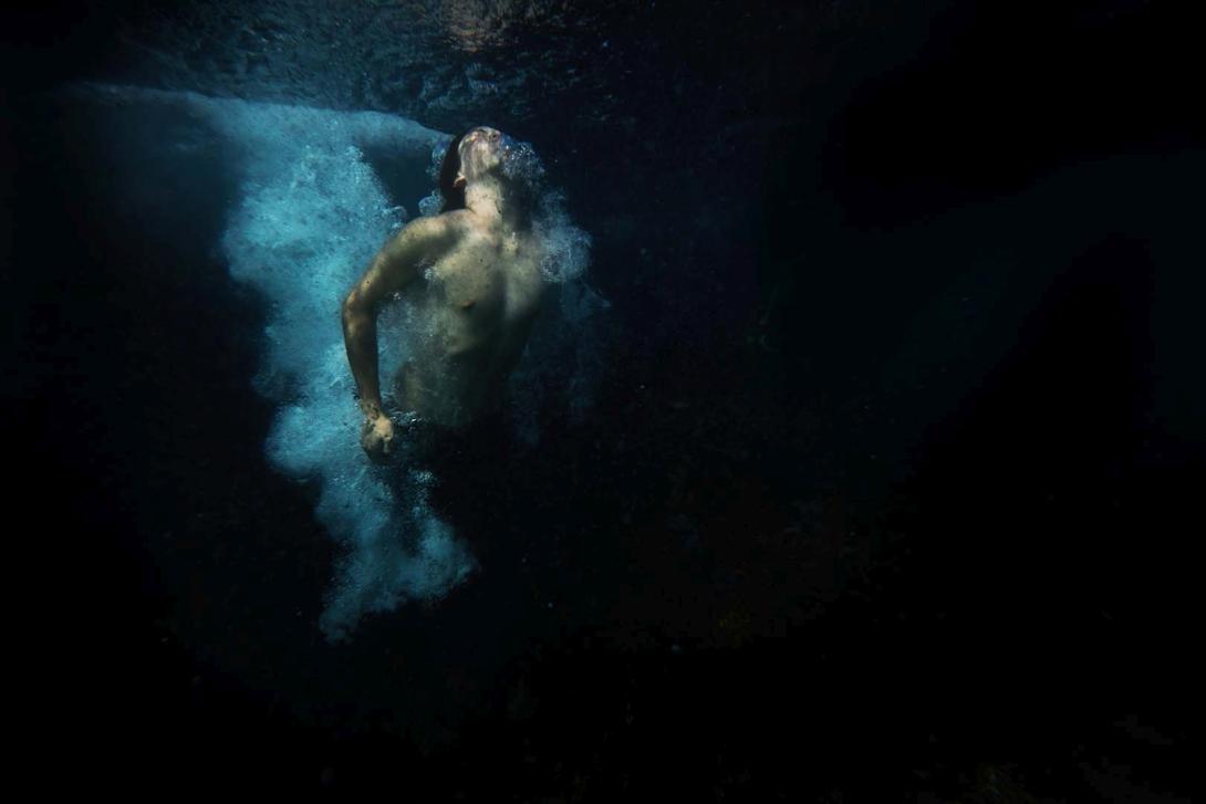 A still photograph of a masculine-appearing person making a splash in dark, almost black water.